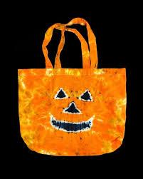 An orange tie-dye tote bag with two handles features a smiling jack-o'-lantern face on the front, set against a black background.