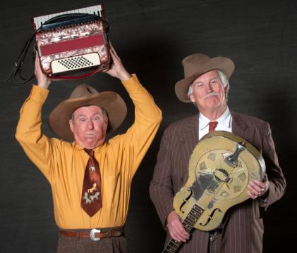Two older men in cowboy hats pose together; one in a yellow shirt and brown tie lifts an accordion above his head, while the other, in a striped suit, holds a guitar. Both have playful expressions and vintage western attire.