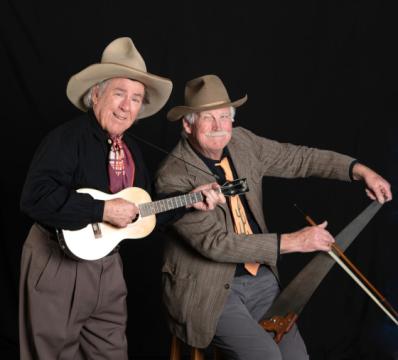 Two older men in cowboy hats pose against a black background. One plays a small guitar or ukulele, smiling, while the other plays a saw with a bow, also smiling. Both wear western-style clothing.
