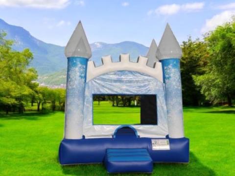 A blue and white inflatable bounce house shaped like a castle stands on green grass in a park, with mountains and trees in the background under a clear blue sky.