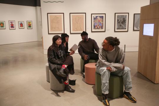 Four people sit and talk in a modern art gallery with framed artwork on the walls behind them. The space has polished floors and bright lighting, and one person is holding a brochure.