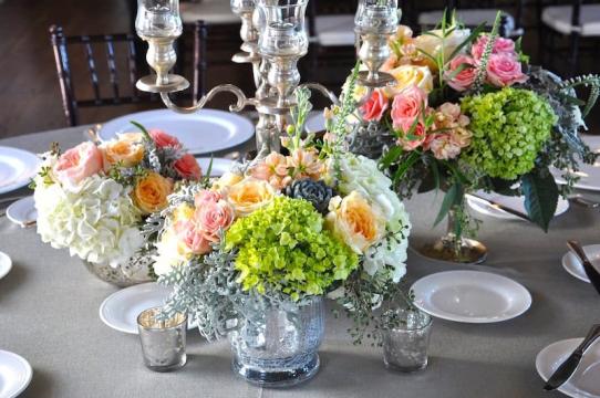 Elegant table setting with vases of colorful flowers, including green hydrangeas, pink and peach roses, white blooms, and greenery, surrounded by plates, glassware, and a silver candelabra centerpiece.