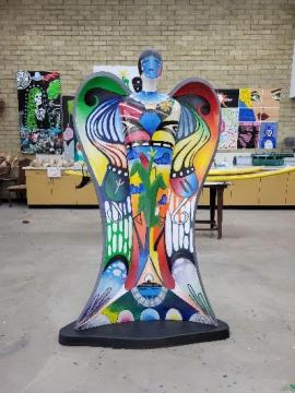 A colorful, abstract angel statue with wings stands in an art classroom. The figure is covered in vibrant, painted patterns and shapes. Artworks and supplies are visible on the walls and tables in the background.