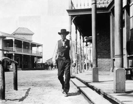 A man in a cowboy hat and vest, wearing a sheriff's badge, confidently walks down a deserted street in a Western town with old wooden buildings and porches.