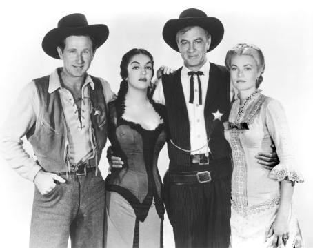 Four people dressed in Old West costumes stand together, two men wearing cowboy hats and vests, one with a sheriff’s badge, and two women in period dresses, all looking at the camera against a plain background.
