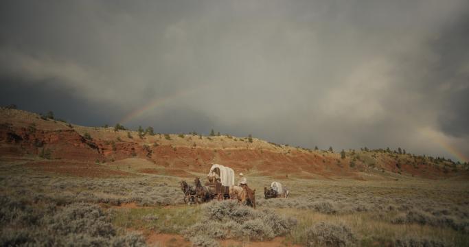 A few covered wagons pulled by horses travel across a grassy plain with red hills in the background, under a cloudy sky with a faint rainbow arching overhead.