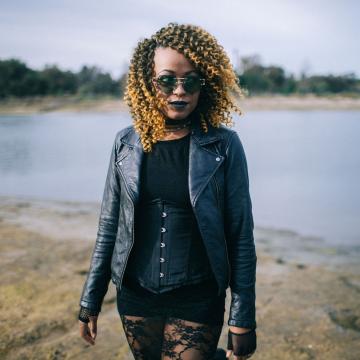 A woman with curly blonde hair, wearing sunglasses, a black leather jacket, black corset, and lace skirt stands outdoors near a body of water, looking confidently at the camera.