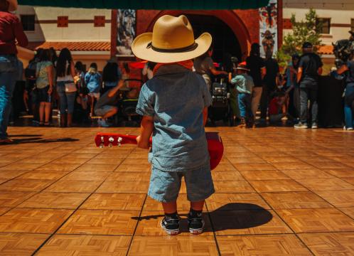 A young child in a cowboy hat and denim outfit stands on a wooden tile floor, holding a red toy guitar, facing a crowd of people gathered in front of a building.
