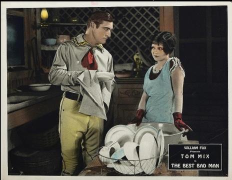 A man in a cowboy outfit dries dishes while talking to a woman in a blue dress and apron. They stand in a rustic kitchen. A sign reads, “William Fox presents Tom Mix in The Best Bad Man.”.