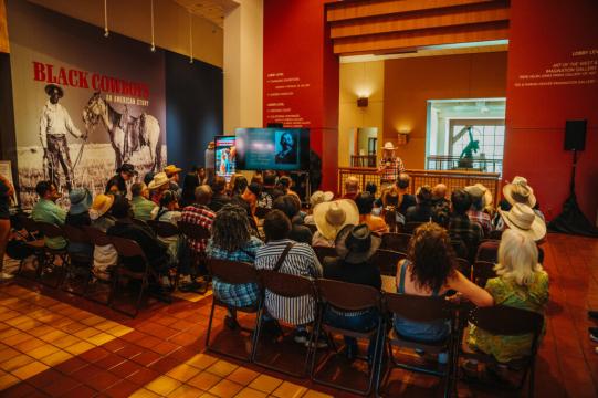 A group of people in cowboy hats sit facing a speaker in a museum exhibit hall with a large “Black Cowboys” sign and historical photos on the wall. The atmosphere is warm and educational.