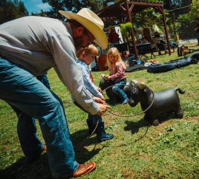 An adult helps a young boy lasso a toy bull while a young girl sits on another toy bull nearby. They are outdoors on grass with people and picnic tables in the background.