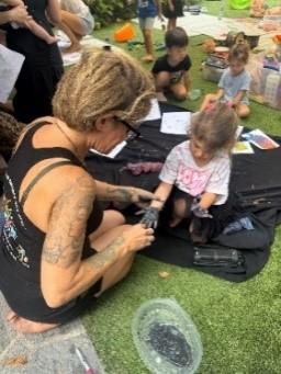 A woman with tattoos helps a young girl with paint-covered hands during an outdoor art activity. Other children sit on the grass nearby, surrounded by art supplies and drawings.