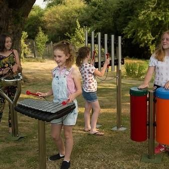 Four girls play various outdoor musical instruments, including drums, chimes, and a xylophone, in a grassy, tree-filled park on a sunny day.