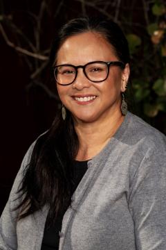 A woman with long dark hair, wearing glasses, earrings, a grey cardigan, and a black top, smiles at the camera with a dark, leafy background behind her.