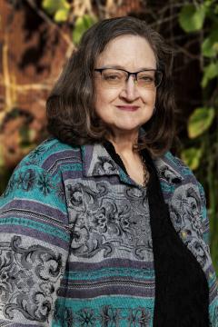 A woman with shoulder-length brown hair and glasses smiles gently while standing outdoors. She is wearing a patterned turquoise, gray, and black jacket over a black top, with green leaves and brown wood in the background.