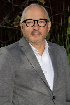 A man with gray hair and a short beard wears black glasses, a gray blazer, and a white shirt, standing outdoors with green plants and brown branches in the background.