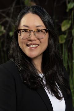 A woman with long dark hair and glasses smiles while wearing a black blazer and white top. The background is blurred greenery.