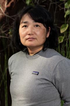 A person with shoulder-length black hair, wearing a grey Patagonia sweatshirt, stands outdoors in front of green plants and wooden fencing, looking slightly past the camera with a neutral expression.