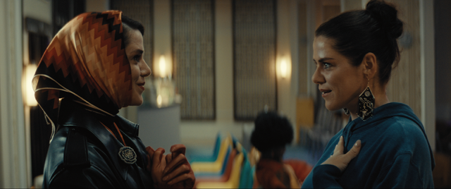 Two women stand facing each other indoors. One wears a colorful headscarf and leather jacket, and the other wears a blue hoodie with large earrings. Both have hands on their chests and appear to be in heartfelt conversation.