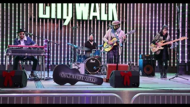 A four-member band performs on an outdoor stage at CityWalk. The musicians play keyboard, drums, electric guitar, and bass. A guitar case labeled “SMILEYBONE JONES” sits in front, and speakers have red bows.