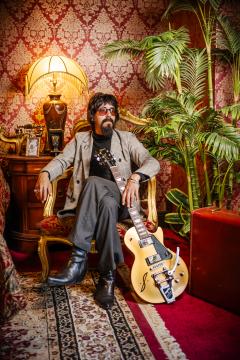 A man with dark hair, sunglasses, and a beard sits in an ornate armchair beside a vintage lamp and lush plants. He wears a gray jacket and black boots, with a cream electric guitar resting next to him.