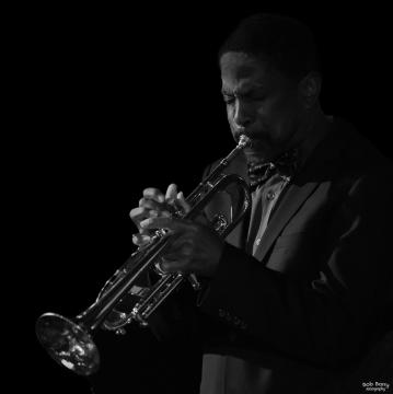 A man in a suit and bow tie plays a trumpet with concentration. The background is black, highlighting the musician and his instrument. The image is in black and white.