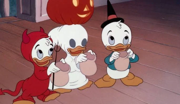 Three cartoon ducklings stand in a row, dressed in Halloween costumes: a devil, a ghost holding a pumpkin lantern, and a witch. Each holds a sack, likely for trick-or-treating, on a wooden floor indoors.