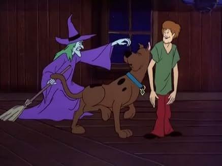 Scooby-Doo and Shaggy look surprised as a green-faced witch in a purple outfit reaches toward them with her hand, standing on a broom inside a wooden, dimly lit room at night.