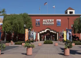 Entrance to the Autry Museum of the American West with banners reading "Indian Country," "Challenge," and "Discover," potted plants, and two flags flying above the red brick building.