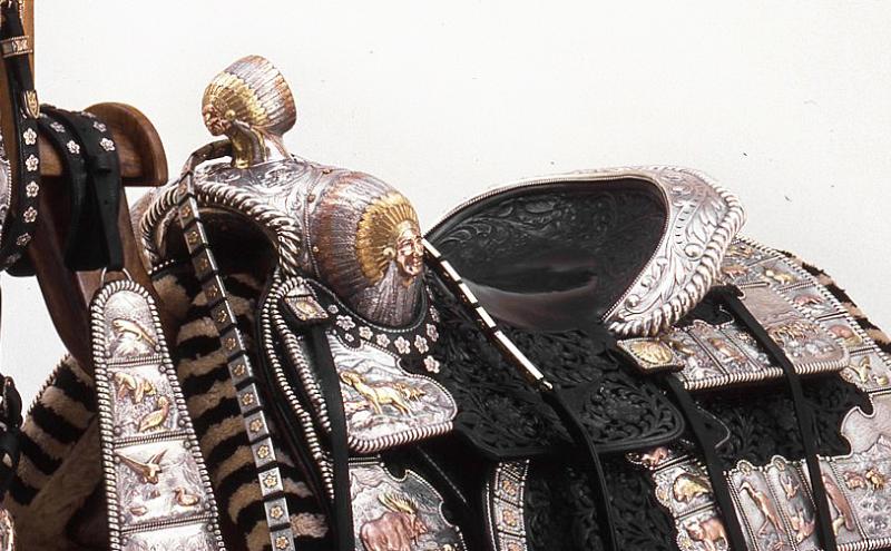 An ornate, Western-style saddle with detailed silver and gold embellishments, intricate leather tooling, and decorative straps, displayed against a plain white background.
