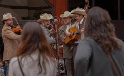 A band dressed in old-fashioned Western attire performs outdoors at the Autry Museum, drawing a crowd. The scene includes musicians playing instruments such as guitars and violins. The museum's "After Hours" event is advertised with a yellow frame on the left side.