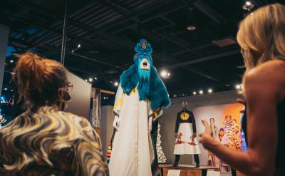Two people observe an elaborate costume on display in a dimly lit exhibition. The costume features vibrant blue fur and an intricate mask with a headdress. Another costume on a mannequin and various colorful outfits are visible in the background.
