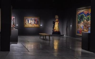 A dimly lit art gallery features colorful paintings on dark walls and a sculpture wearing a golden costume with a headdress. A wooden bench is placed on a polished concrete floor, providing a spot for visitors to sit and admire the artwork.