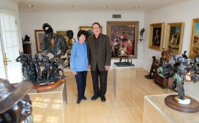 A couple stands in an art gallery filled with sculptures and paintings. They are surrounded by various artworks, including bronze sculptures and framed paintings on the walls. The room has a bright, inviting atmosphere.