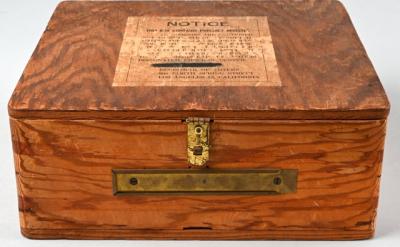 A vintage wooden box with a brass latch and a rectangular brass label holder. The top has a printed notice with text in capital letters. The wood has a textured grain and a warm, natural color.