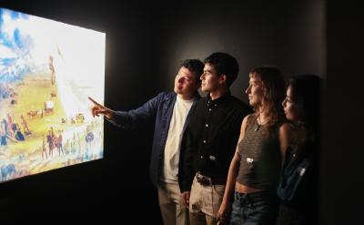 Four people stand in a dimly lit room, attentively viewing and discussing a large, illuminated painting on the wall. One person points toward the artwork, as the others look on with interest.