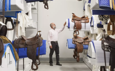 A man in a white shirt and black pants examines intricately designed saddles in a storage room. The saddles are mounted on racks, arranged on blue and white protective padding. The room has shelves and a bright atmosphere.