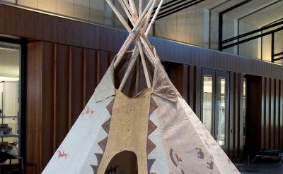 A traditional tipi with painted designs stands indoors on a modern floor, surrounded by wood-paneled walls and large windows in a spacious, contemporary building.