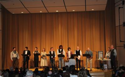 A group of people stand on a stage in front of a beige curtain, some holding scripts. An audience is seated in the foreground, watching the performers. A person stands in front of the stage, possibly directing or addressing the group.