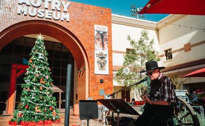 A musician in a cowboy hat plays guitar and sings outside the Autry Museum, near a large decorated Christmas tree. The scene is sunny, with festive holiday decorations and museum banners visible.
