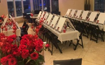 A room is set up for a painting class with rows of easels and blank canvases on tables, each with a red cup. In the foreground, a bouquet of red roses adds a decorative touch. Large windows are visible in the background.