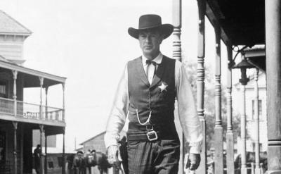 A sheriff wearing a vest, cowboy hat, and star badge walks confidently down a deserted street in a classic Western town with wooden buildings and porches in the background.