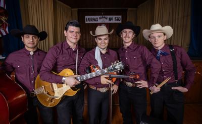 Five men in matching maroon shirts and cowboy hats pose on stage with musical instruments, including a guitar, upright bass, violin, and drumsticks. A sign reading “Fairfax Grange No 570” hangs in the background.
