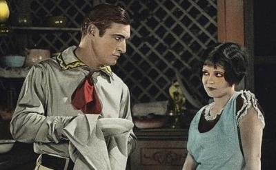 A man in a western-style shirt and red neckerchief dries a dish while looking at a woman in a blue dress, who gazes back with a skeptical expression. They stand in a rustic kitchen with shelves and dishes behind them.