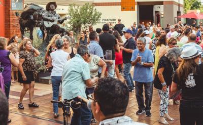 A lively outdoor event with a diverse crowd dancing and enjoying live music; a musician plays guitar on stage, and people of all ages fill the plaza under warm daylight.