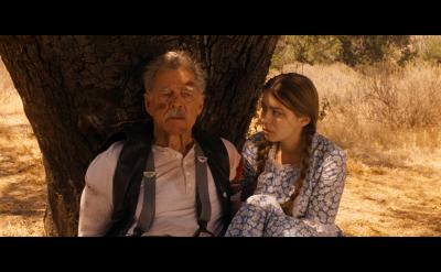 An older man and a young woman sit under a tree in a sunlit field. The man looks ahead seriously, while the woman, in a blue floral dress, gazes at him with concern.