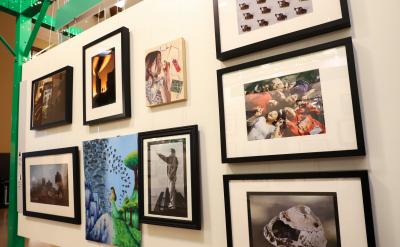 A gallery wall displays various framed artworks, including photographs, illustrations, and paintings, arranged in a grid on a white wall, with part of a green metal beam visible on the left.