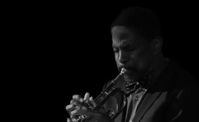 A man in a suit and bow tie plays a trumpet with concentration. The background is black, highlighting the musician and his instrument. The image is in black and white.