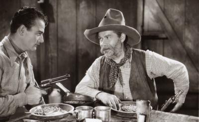 Two men in Western attire sit at a table with plates of food. One man points a revolver while the other, wearing a cowboy hat and scarf, looks at him. The setting appears to be an old wooden cabin.