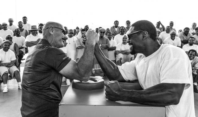 two inmates arm wrestle, their muscles are bulging, and an enthusiastic crowd of prisoners are gathered behind them crowd 